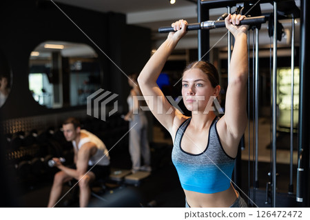 Slender athletic girl work out muscles of arms and shoulders on simulator in gym of fitness club 126472472