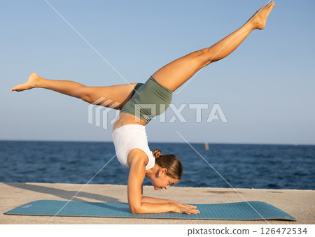 On windy day young slender woman does yoga near sea doing pincha mayurasana 126472534