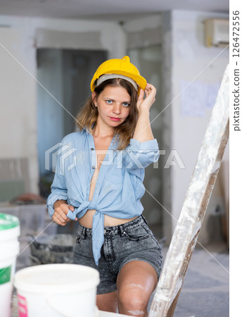 Seductive woman in hardhat and denim shirt posing next to a stepladder in room being renovated 126472556