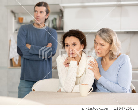 Man quarrels with his wife in kitchen, standing behind her, and elderly mother hugs and calms her daughter 126472593