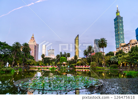 Evening view of Taipei modern skyline in Taiwan with skyscrapers rising above the cityscape, reflected in the tranquil pond of Sun Yat Sen Memorial Hall Park, surrounded by lotus plants and palm trees 126475618