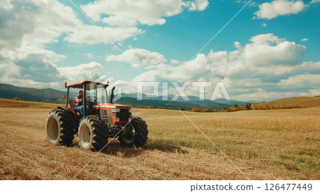 A farmer operates a red tractor across a vast harvested field, surrounded by rolling hills and a cloudy blue sky, embodying the essence of modern agriculture 126477449