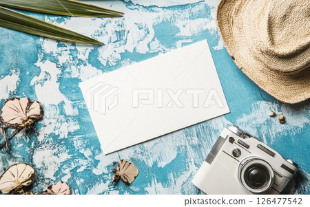 Tropical travel flat lay featuring blank card, straw hat, camera, and palm leaves on rustic blue background, perfect for vacation planning or memories 126477542