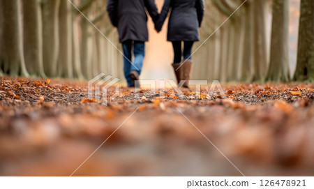 Couple walking hand in hand along a tree-lined path covered with autumn leaves, surrounded by tall trees, creating a serene and romantic atmosphere in a beautiful outdoor setting 126478921