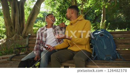 Two happy biracial male friends sitting and using smartphone in park with backpacks Two happy biracial male friends sitting and using smartphone in park with backpacks 126482378