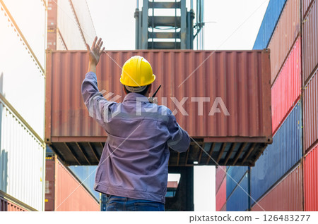 Dock supervisor in yellow hard hat and uniform directing container loading with walkie talkie and hand gesture at shipping container yard. 126483277