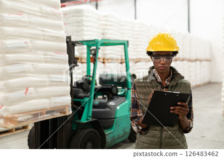 Warehouse worker wearing safety gear checking inventory with clipboard near forklift 126483462