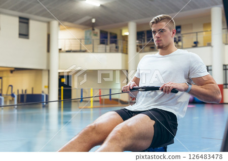 Focused man using rowing machine in sports center, building strength and endurance, copy space 126483478