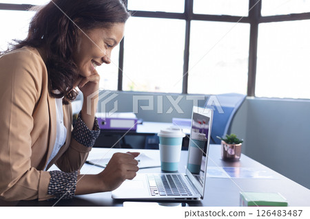 Smiling woman using laptop at office desk with coffee cup nearby 126483487