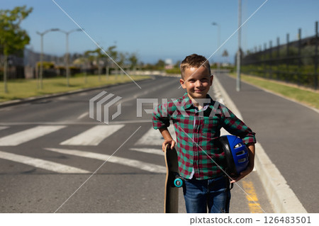 Smiling boy holding skateboard and helmet standing on school road, copy space 126483501