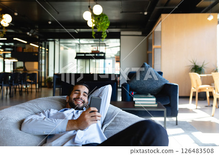 Relaxing in modern office, man smiling while using smartphone on bean bag 126483508