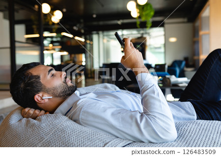 Relaxing in office lounge, man using smartphone and wearing wireless earbuds Relaxing in office lounge, man using smartphone and wearing wireless earbuds 126483509