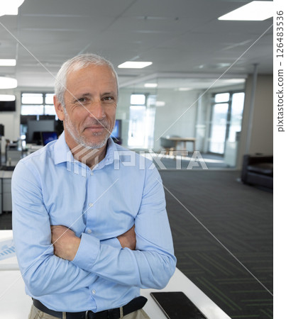 Senior businessman standing with arms crossed in modern office, wearing blue shirt Senior businessman standing with arms crossed in modern office, wearing blue shirt 126483536