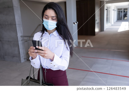 Businesswoman wearing mask texting on smartphone while walking in office hallway, copy space Businesswoman wearing mask texting on smartphone while walking in office hallway, copy space 126483549
