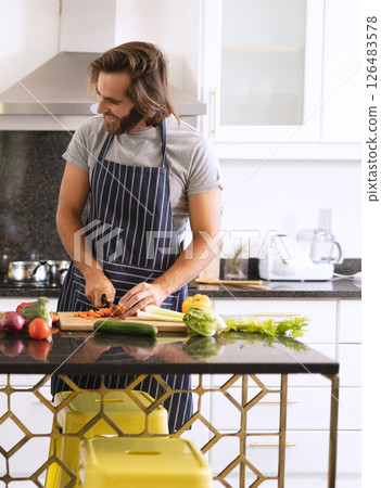 Man in kitchen chopping vegetables on counter, wearing apron, smiling while cooking 126483578