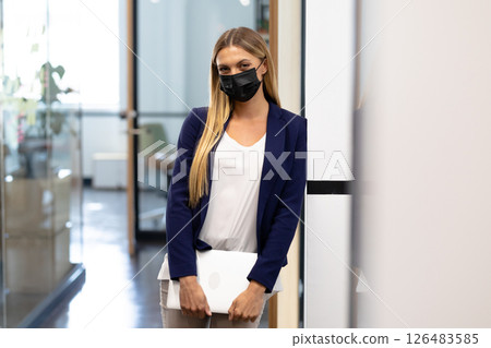 Professional woman in office holding laptop, wearing mask, looking confident 126483585