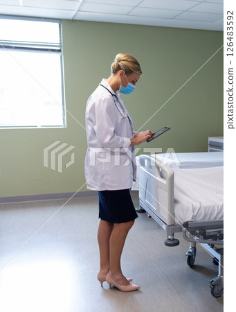 In hospital, doctor wearing mask reviewing patient information on digital tablet 126483592