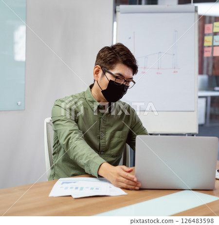 Asian man wearing mask using laptop in office, analyzing business charts Asian man wearing mask using laptop in office, analyzing business charts 126483598