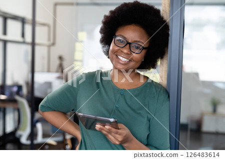 Smiling woman in office holding tablet, leaning against glass wall confidently 126483614