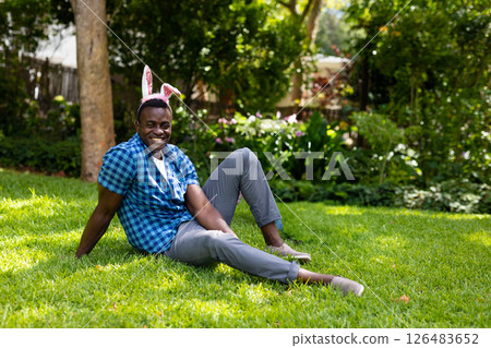 Smiling man wearing bunny ears sitting on grass in sunny garden 126483652
