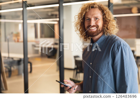 Smiling man with smartphone standing in modern office, looking confident, copy space 126483696