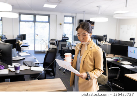 Businesswoman reviewing documents while holding coffee cup in modern office, copy space 126483727
