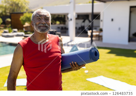 Elderly man holding yoga mat, smiling outdoors near home pool, enjoying sunshine, copy space 126483735