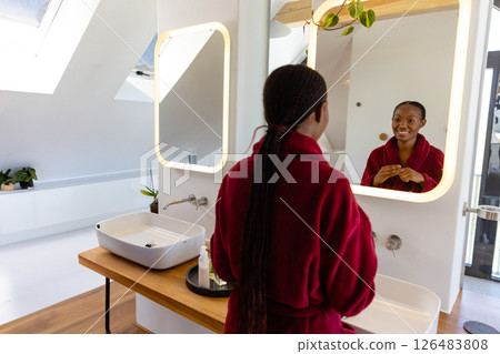 Woman in red robe smiling at mirror in modern bathroom, enjoying morning routine 126483808