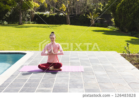 Woman meditating on yoga mat by pool in peaceful outdoor garden setting, copy space 126483828