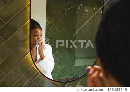 Woman in white robe brushing teeth in modern bathroom, reflecting in mirror 126483849