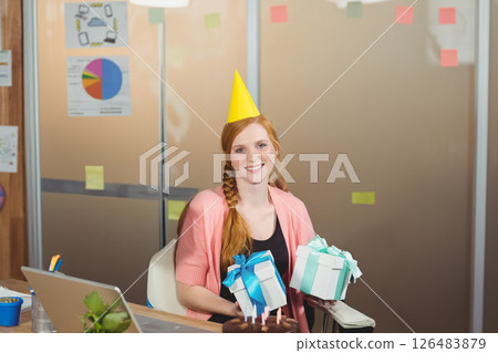 Smiling woman in party hat holding gifts at office birthday celebration 126483879