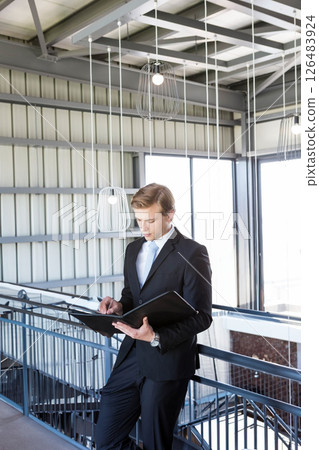 Businessman in suit reviewing documents in modern office building hallway Businessman in suit reviewing documents in modern office building hallway 126483924