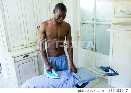 Shirtless man ironing clothes in kitchen, smiling and enjoying household chores 126483947