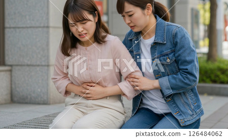 A woman sitting down in the street with stomach pain and a woman caring for her A woman sitting down in the street with stomach pain and a woman caring for her 126484062