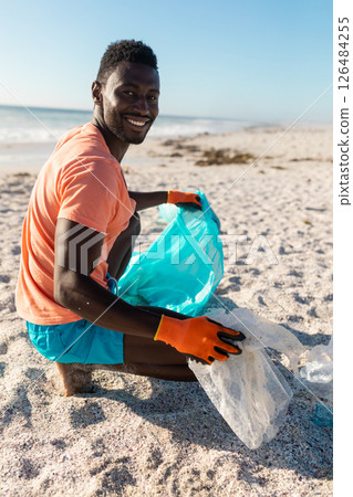 Smiling man cleaning beach, collecting plastic waste with blue bag, sunny day, copy space 126484255