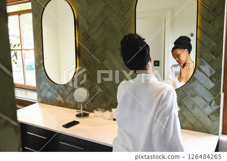 Woman in white robe applying skincare in bathroom, reflecting in mirror, copy space 126484265