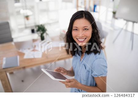 Smiling Asian woman using tablet in bright office, surrounded by documents, copy space 126484317