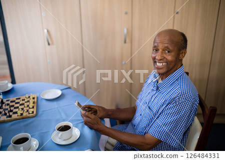 Elderly man smiling while using smartphone at table with coffee and chessboard, copy space 126484331
