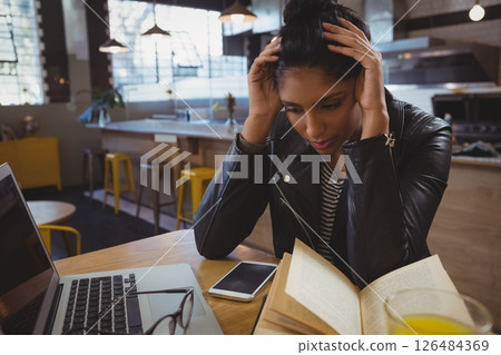 Young woman in cafe reading book, looking stressed, laptop and smartphone nearby 126484369