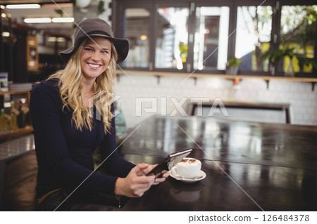 Smiling woman in hat using tablet at cafe with cappuccino on table, copy space 126484378