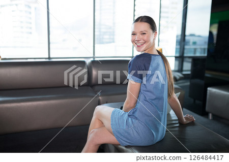 Smiling woman in modern office wearing casual dress, sitting on sofa, copy space 126484417
