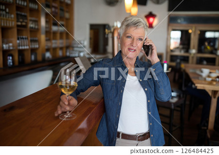Mature woman enjoying wine while talking on smartphone at cozy bar, copy space Mature woman enjoying wine while talking on smartphone at cozy bar, copy space 126484425