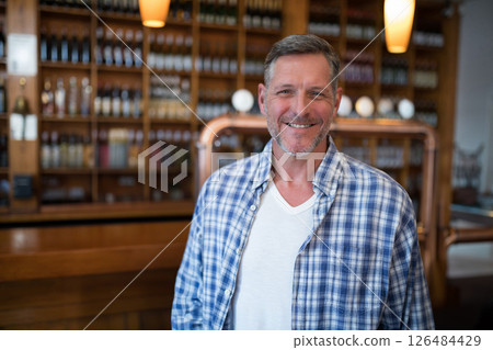 Smiling man in plaid shirt standing at bar with wine bottles in background, copy space 126484429