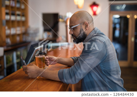 Man enjoying beer while browsing smartphone at cozy bar counter, copy space Man enjoying beer while browsing smartphone at cozy bar counter, copy space 126484430