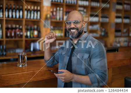 Smiling man at bar holding smartphone, enjoying drink with shelves of bottles, copy space Smiling man at bar holding smartphone, enjoying drink with shelves of bottles, copy space 126484434