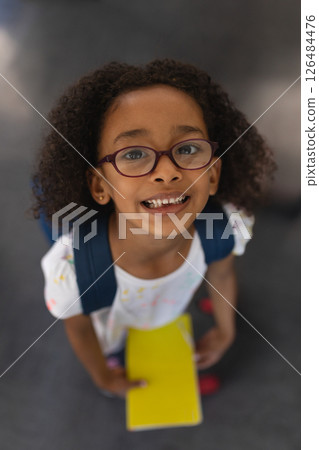 Smiling girl with backpack holding yellow notebook, looking up in school hallway 126484476