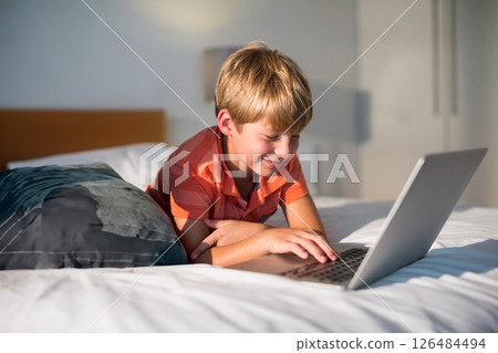 Young boy using laptop on bed at home, smiling and enjoying online activity 126484494