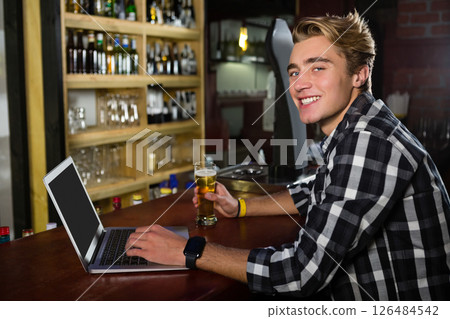 Young man using laptop and enjoying beer at bar, smiling at camera, copy space 126484542