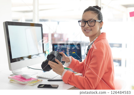 Asian woman holding camera, smiling at desk with computer and smartphone, copy space 126484583