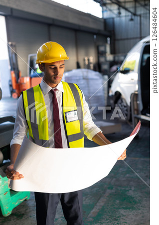 Engineer in hard hat reviewing blueprint in busy warehouse environment 126484604